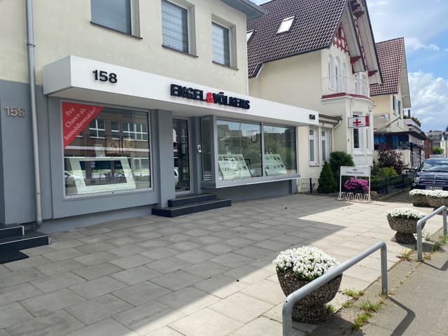 Exterior of Engel & Völkers real estate office at 158, with large windows displaying property listings. Flower pots line the sidewalk.