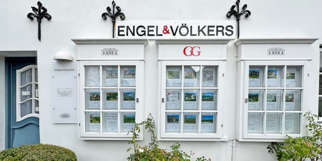 Engel & Völkers Immobilienbüro mit weißer Fassade, Auslagen in den Fenstern und blauer Tür auf der linken Seite.