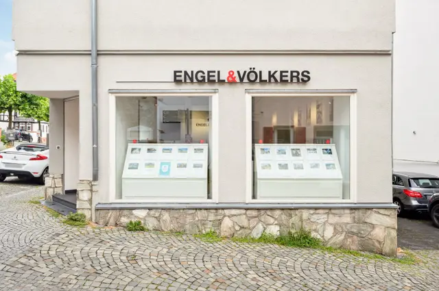 Exterior of Engel & Völkers real estate office with property listings displayed in the windows. Cobblestone street in front.