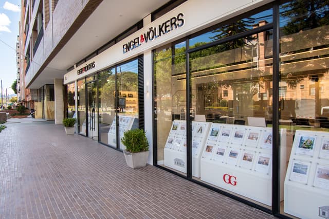 Engel & Völkers real estate office with large glass windows displaying property listings on white stands. A brick sidewalk leads to the entrance.