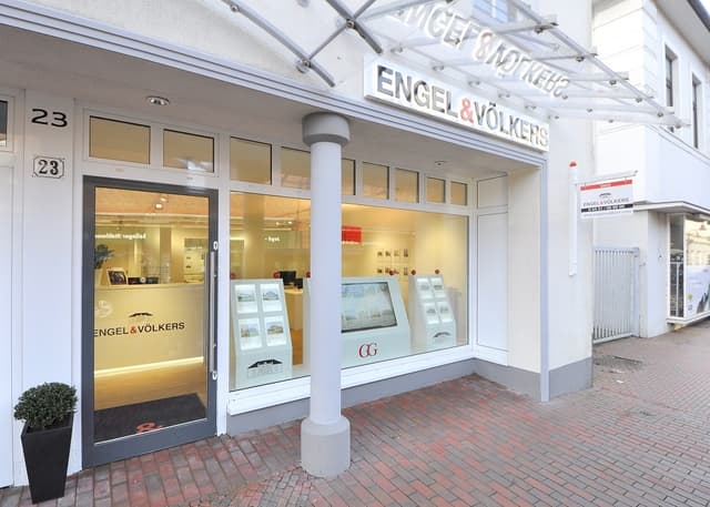 Engel & Völkers real estate office exterior. White building with glass windows displaying property listings. Red brick sidewalk.