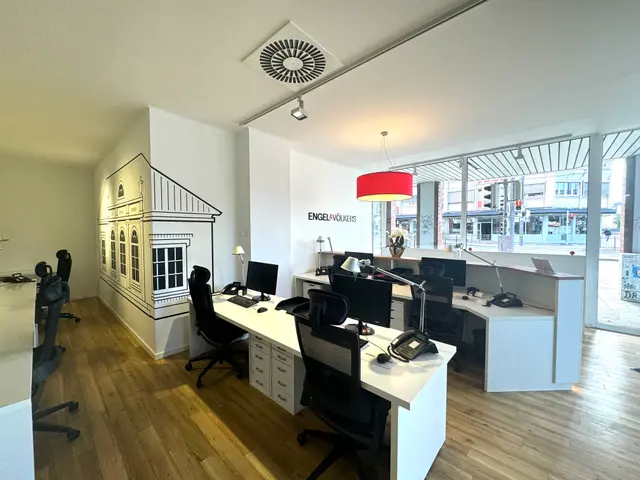Engel & Völkers office interior with white desks, black chairs, computers, and a building mural on the wall. A red lamp hangs from the ceiling.