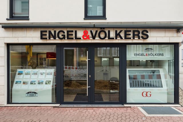 Exterior of Engel & Völkers real estate office with black framed windows and doors. A display of property listings is visible.