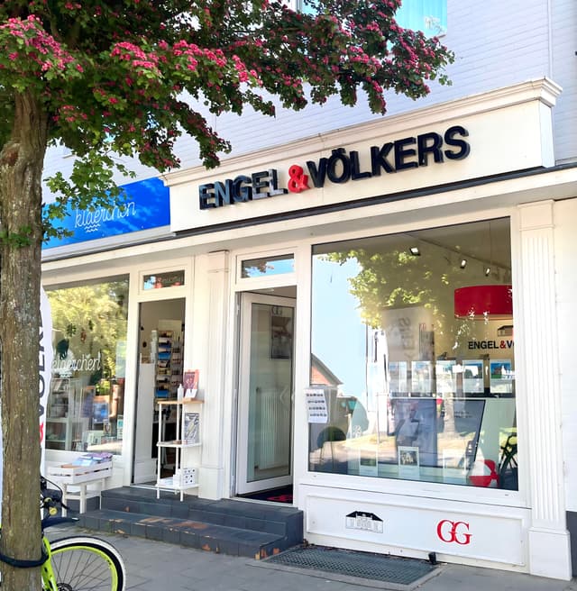 Engel & Völkers real estate office storefront with white trim and black lettering. A tree with pink flowers is on the left.