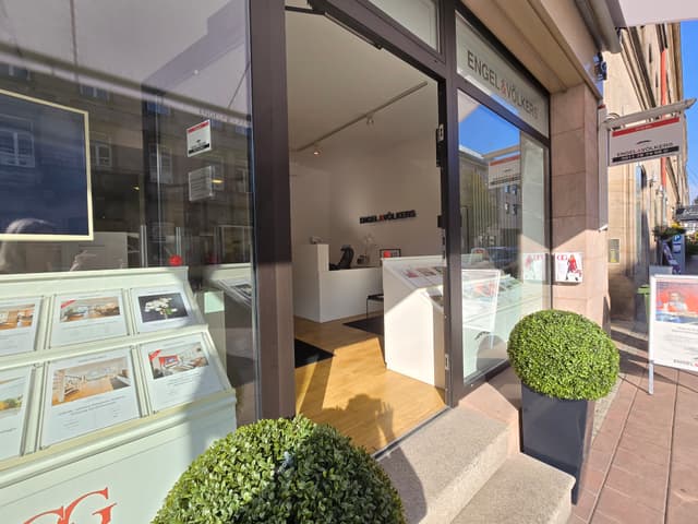 Engel & Völkers Immobilienbüro mit offener Glasfront. Innen sind ein weißer Empfangstresen und Immobilienangebote sichtbar. Grüne Formschnittpflanzen flankieren den Eingang.