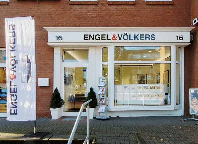 Engel & Völkers Immobilienbüro mit einer Backsteinfassade. Eine weiße Flagge mit dem Firmenlogo befindet sich auf der linken Seite. Die Adresse "16" befindet sich über dem Eingang.